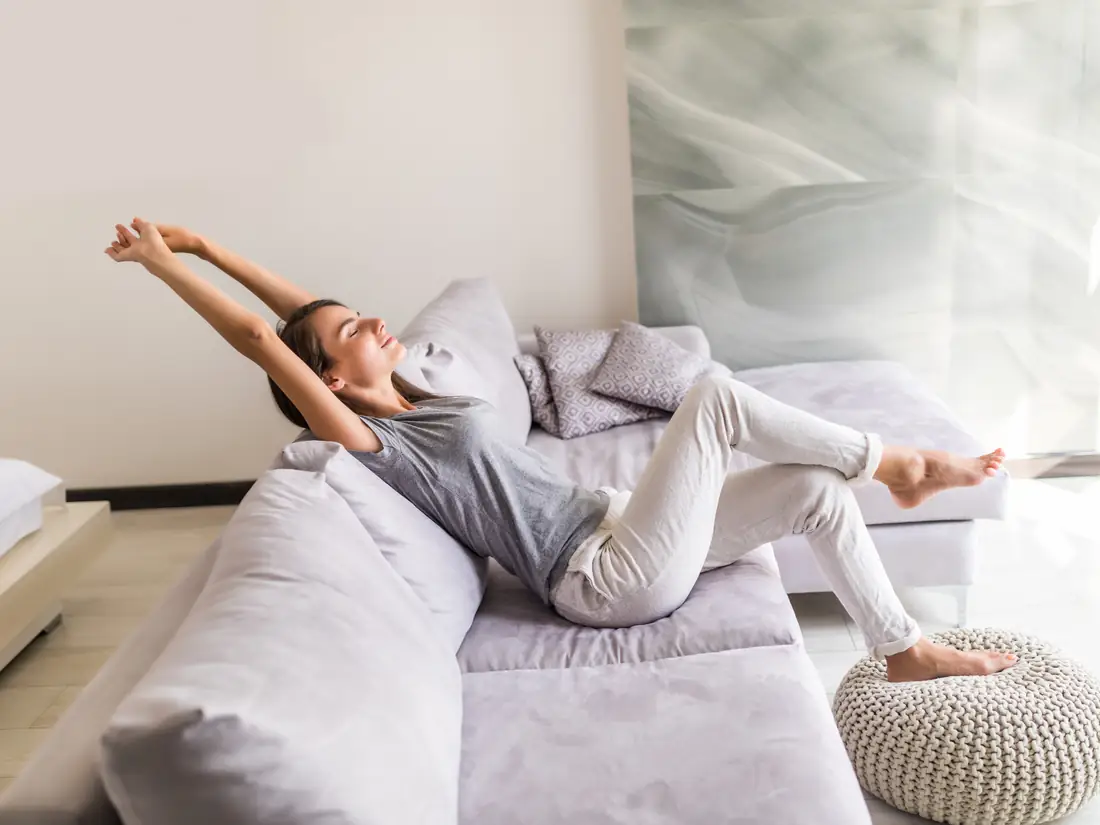 Woman stretching contentedly on a pale grey couch in a bright, cool living room